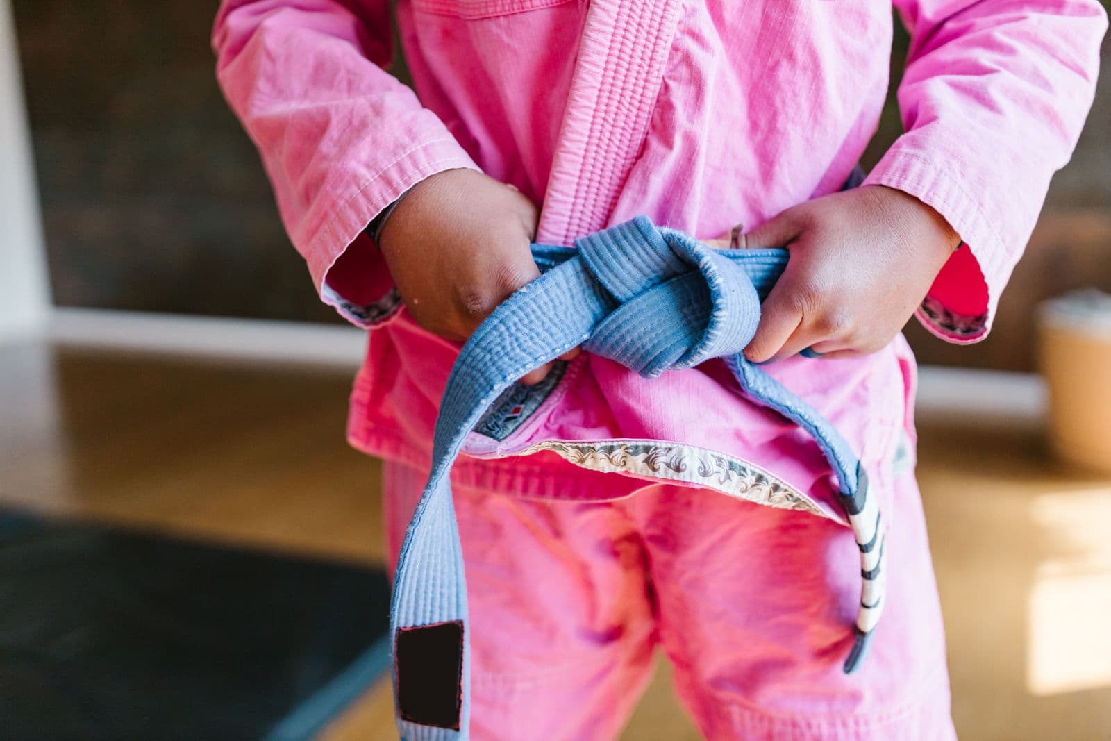 Un enfant en tenue d'arts martiaux rose qui noue une ceinture bleue, vu de la poitrine aux genoux