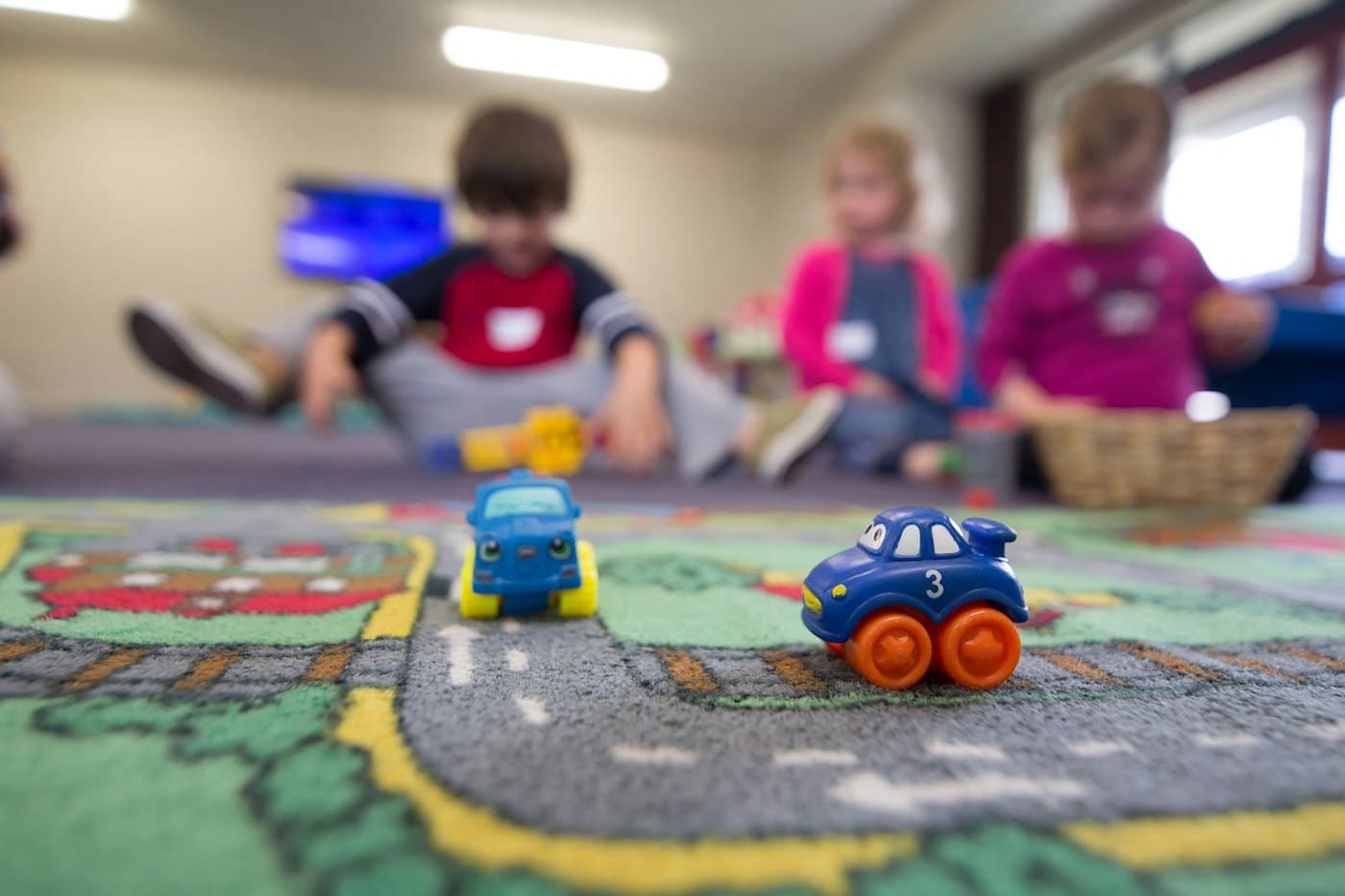 Des petites voitures sur un tapis de jeu, avec des enfants qui jouent à l'arrière-plan