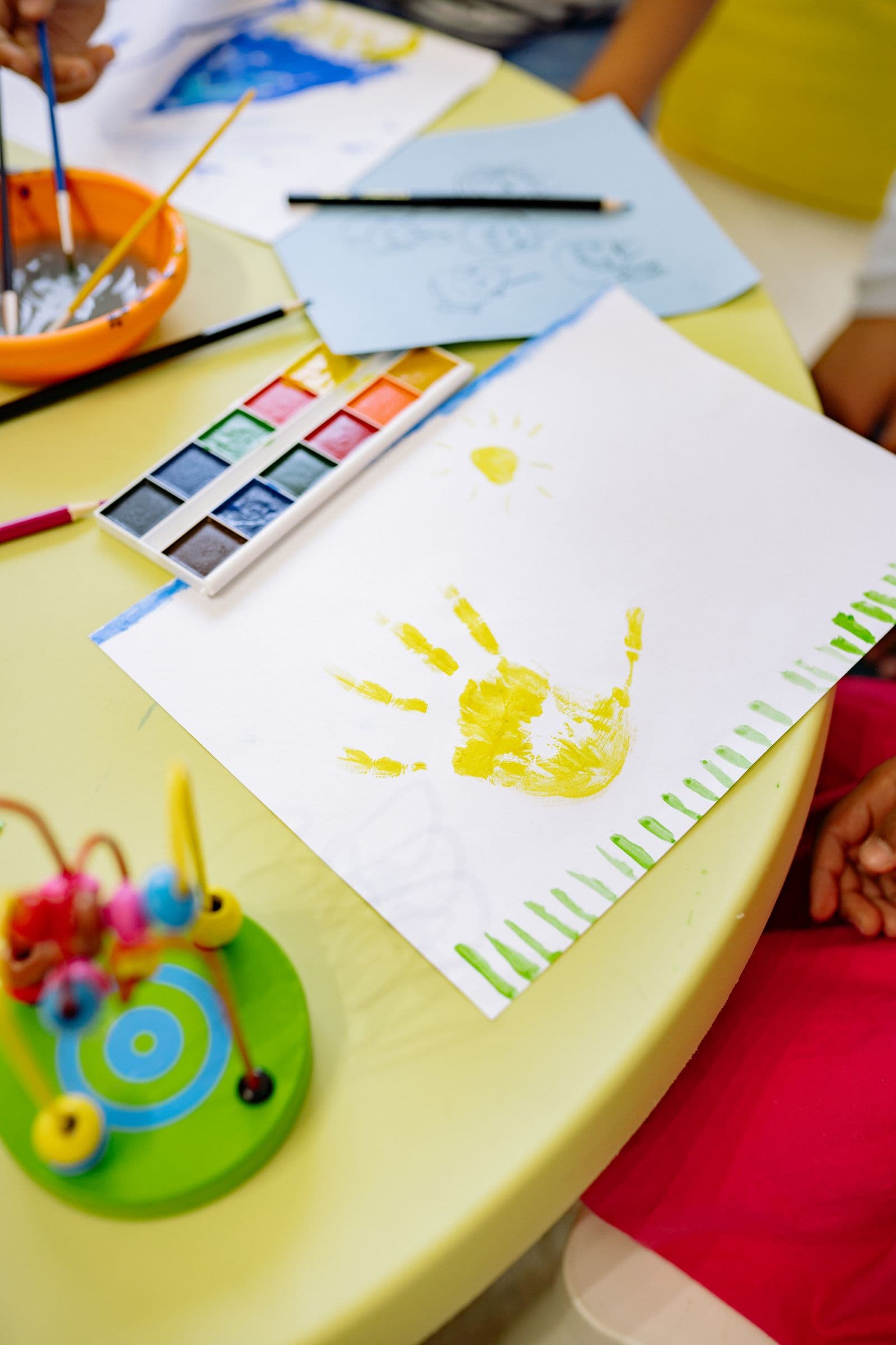 Une empreinte de main jaune peinte à côté d'aquarelles et d'un jouet à perles sur une table de prématernelle