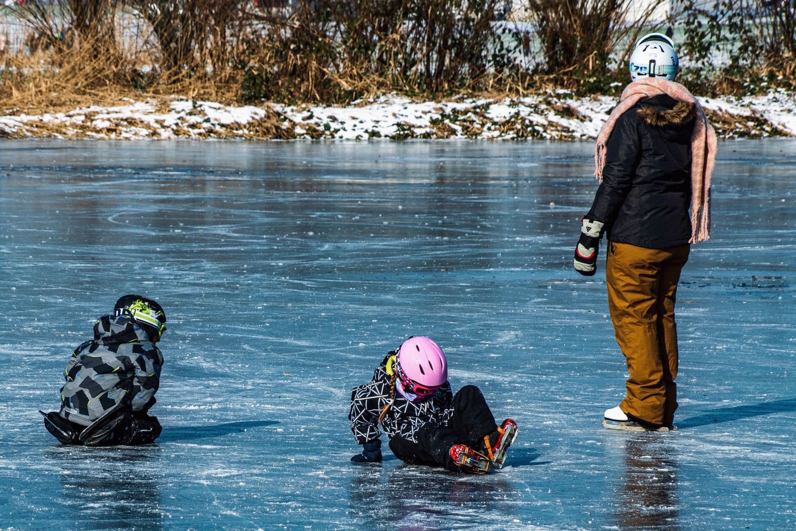 Deux jeunes enfants casqués et en patins sur la glace, avec un adulte qui patine à côté, vus de dos